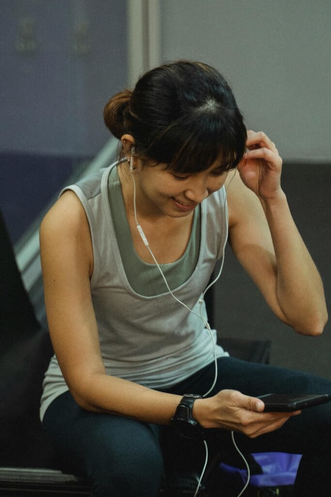 Smiling Asian woman using smartphone and earphones during gym break, enjoying a video call.